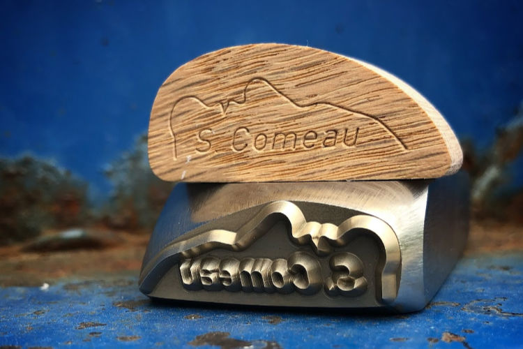 end grain wood shows s comeau logo stamped on it and the steel stamp made by Buckeye engraving lies under the piece of wood showing the deeply engraved sharp face on the steel wood hand stamp